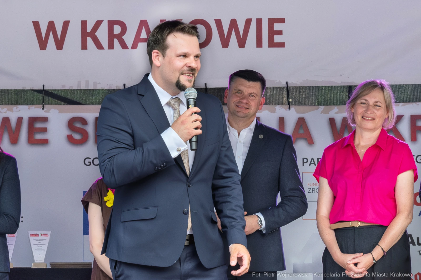 Mężczyzna w garniturze przemawia na scenie, obok niego stoją dwie osoby.  Autor: Piotr Wojnarowski