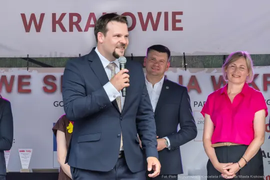 Mężczyzna w garniturze przemawia na scenie, obok niego stoją dwie osoby.