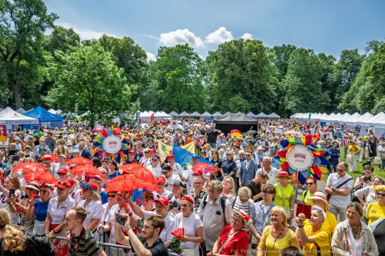 Tłum ludzi na festiwalu, kolorowe parasole, stoiska i zielone drzewa w tle.