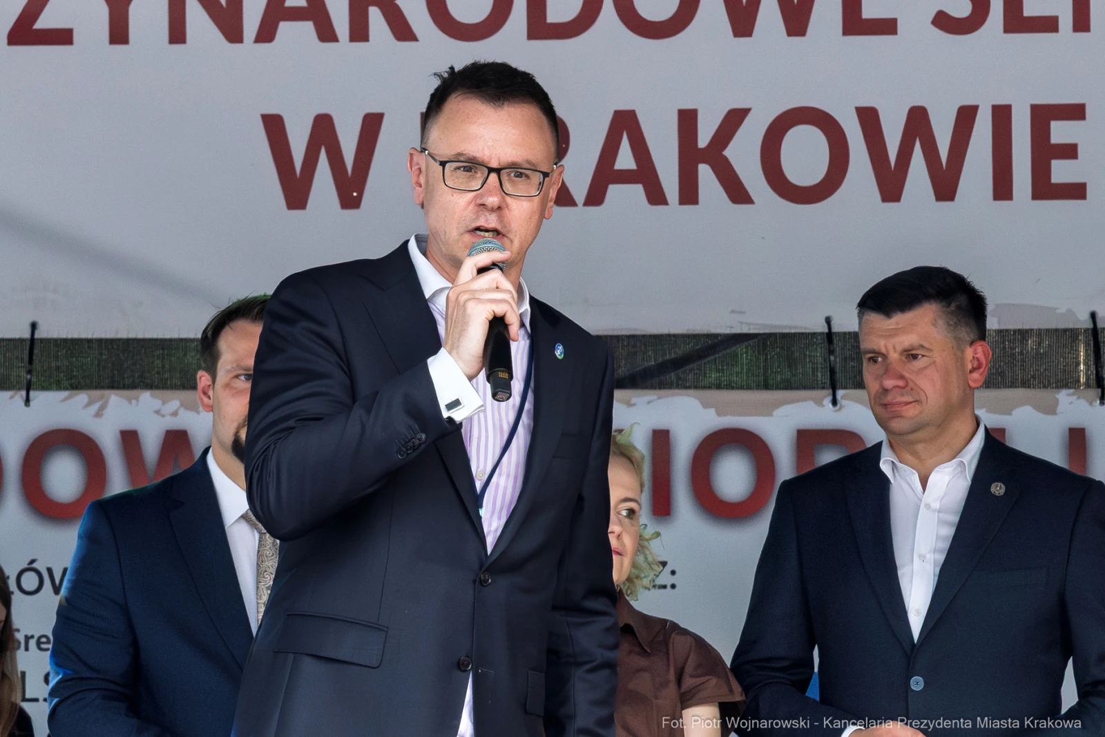 Mężczyzna w garniturze przemawia do mikrofonu na tle banera z napisem o wydarzeniu w Krakowie.  Autor: Piotr Wojnarowski