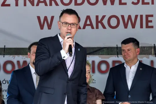 Mężczyzna w garniturze przemawia do mikrofonu na tle banera z napisem o wydarzeniu w Krakowie.