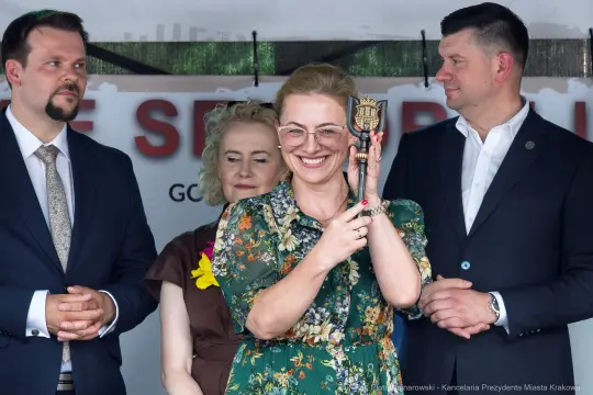 Kobieta w kwiecistej sukience trzyma nagrodę, obok niej stoją mężczyźni i kobieta w tle.
