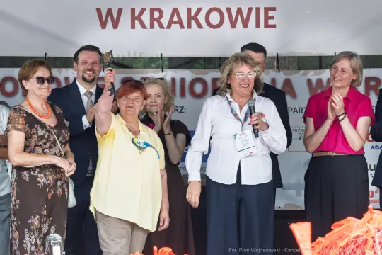 Uczestnicy wydarzenia w Krakowie, kobieta trzymająca dzwonek, aplauz i uśmiechy.