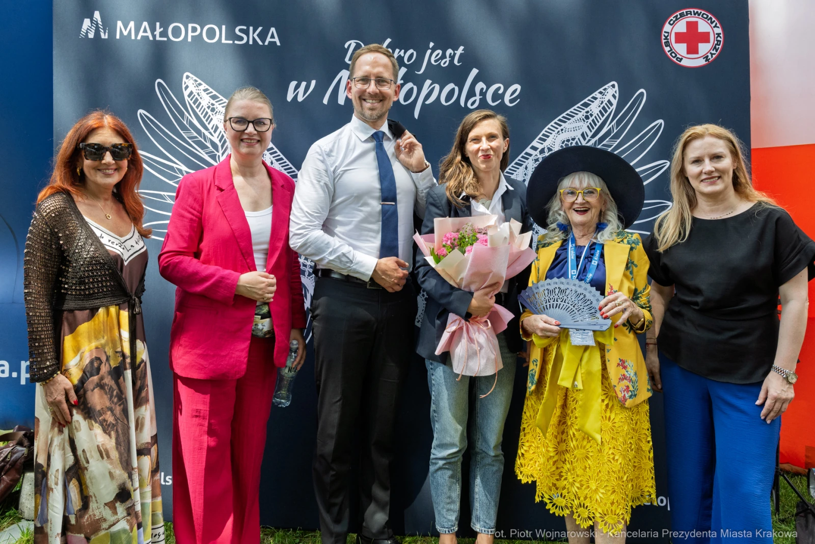 Grupa osób pozujących na tle baneru promującego Małopolskę, z kwiatami i uśmiechami.  Autor: Piotr Wojnarowski