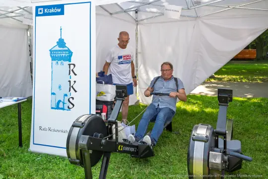 Mężczyzna ćwiczy na ergometrze wioślarskim, obok stoi inny mężczyzna. W tle baner z logo RKS.
