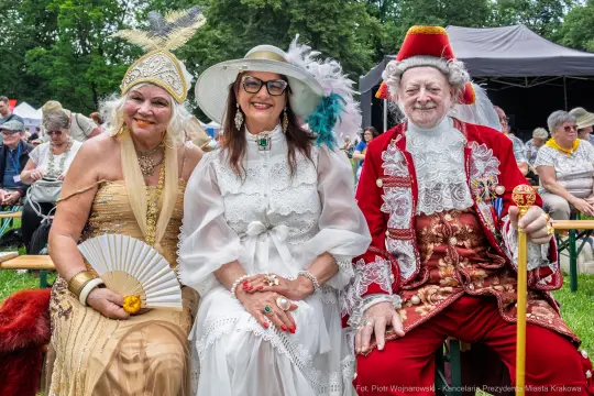 Trzy osoby w eleganckich strojach, uśmiechające się, w plenerze podczas wydarzenia kulturalnego.