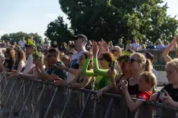 Tłum ludzi na koncercie, niektórzy w koronach z kwiatów, klaskają i cieszą się muzyką.