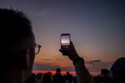 Osoba trzymająca telefon, nagrywająca zachód słońca w tłumie ludzi.