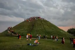 Ludzie wspinający się na zieloną górę, w tle chmury i trawa.