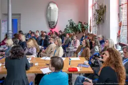 Grupa ludzi uczestniczy w konferencji w jasnym wnętrzu z dużym lustrem i roślinami.