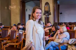 Kobieta w jasnym żakiecie uśmiecha się, w tle grupa osób w sali.