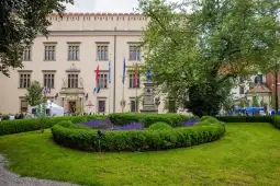 Budynek z flagami, otoczony zielonym ogrodem i kwiatami.