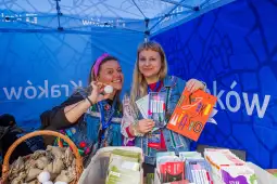 Dwie kobiety w strojach festiwalowych trzymają książki i uśmiechają się przy stoisku.