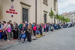 Ludzie stoją w długiej kolejce na ulicy, w tle budynki i drzewa.