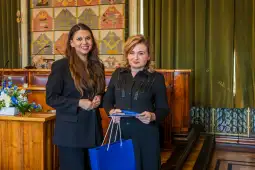 Dwie kobiety w eleganckich strojach, stojące w sali z drewnianymi elementami, trzymają torbę i dokumenty.