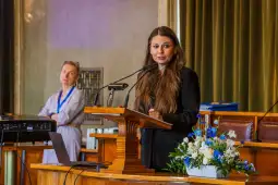 Kobieta przemawia z mównicy, w tle druga osoba obserwuje wydarzenie. Na stole kwiaty.