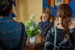 Mężczyzna w eleganckim ubraniu siedzi przy biurku, otoczony przez kwiaty i osoby z tyłu.