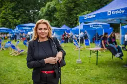 Kobieta w czarnym stroju z czerwoną paskiem na tle namiotów i ludzi w parku.