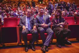 Trzech mężczyzn siedzących na czerwonych fotelach wśród publiczności na wydarzeniu.