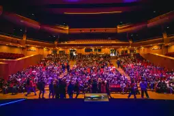 Tłum ludzi w sali teatralnej, przygotowujący się do wydarzenia.