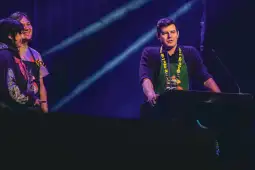 Mężczyzna przemawia z podium, obok niego dwie osoby, w tle kolorowe światła.