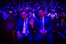 Dwaj mężczyźni w garniturach siedzą na widowni, otoczeni przez tłum ludzi w ciemnej sali.