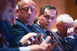 Grupa mężczyzn w garniturach siedzących obok siebie, skupionych na swoich telefonach.