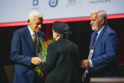 Wręczenie nagrody podczas uroczystości, z osobami trzymającymi kwiaty i statuetkę.