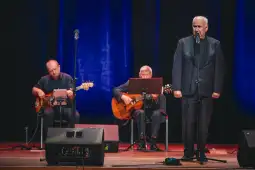 Mężczyzna śpiewa na scenie, towarzyszą mu muzycy grający na gitarach.