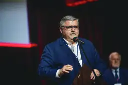 Mężczyzna w okularach przemawia do mikrofonu na tle sceny.