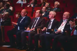 Czterech mężczyzn w garniturach siedzi na widowni, uczestnicząc w wydarzeniu.
