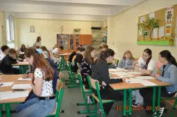 Uczniowie pracują w grupach w klasie, przy biurkach z materiałami edukacyjnymi.