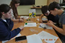 Cztery osoby pracują nad projektem edukacyjnym przy stole, używając materiałów i narzędzi.