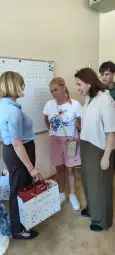 Spotkanie grupy osób w sali, jedna z kobiet trzyma torbę z darami.