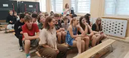 Grupa uczniów siedzących na ławkach w sali, niektórzy trzymają telefony.