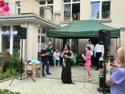 Kobieta w czarnej sukni przemawia na wydarzeniu plenerowym, w tle goście i dekoracje.