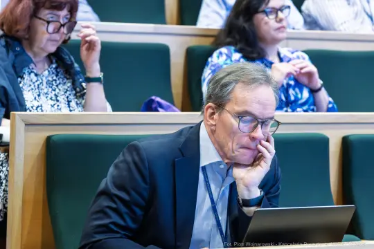 Mężczyzna w garniturze korzysta z laptopa, w tle kobiety siedzące na wykładzie.