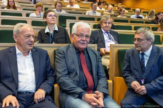 Trzech mężczyzn w garniturach siedzi na widowni w sali konferencyjnej.