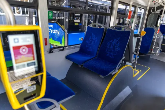 Puste siedzenia w autobusie z niebieskimi tapicerkami i widokiem na zewnętrzny przystanek.