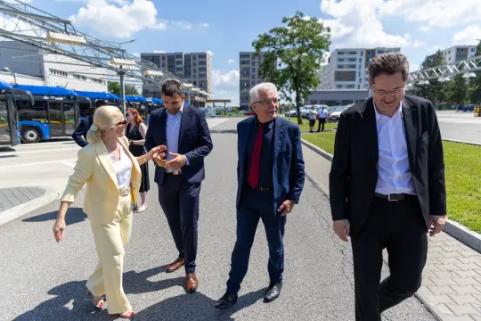 Cztery osoby w eleganckich strojach spacerują po terenie zakładu, w tle nowoczesne budynki.