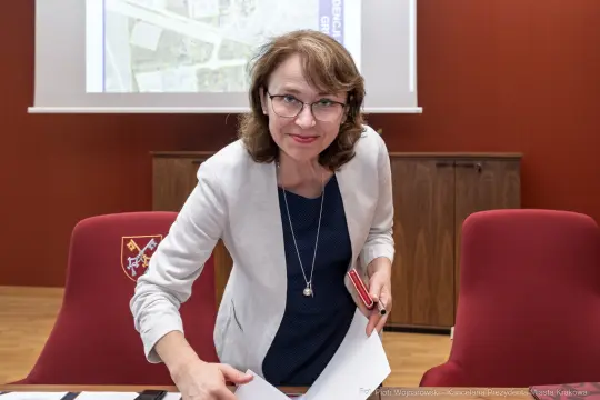 Kobieta w okularach uśmiecha się, przygotowując dokumenty na stole w sali konferencyjnej.