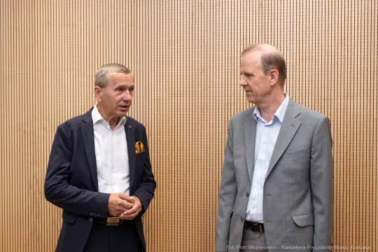 Dwóch mężczyzn rozmawia w sali z drewnianą ścianą.