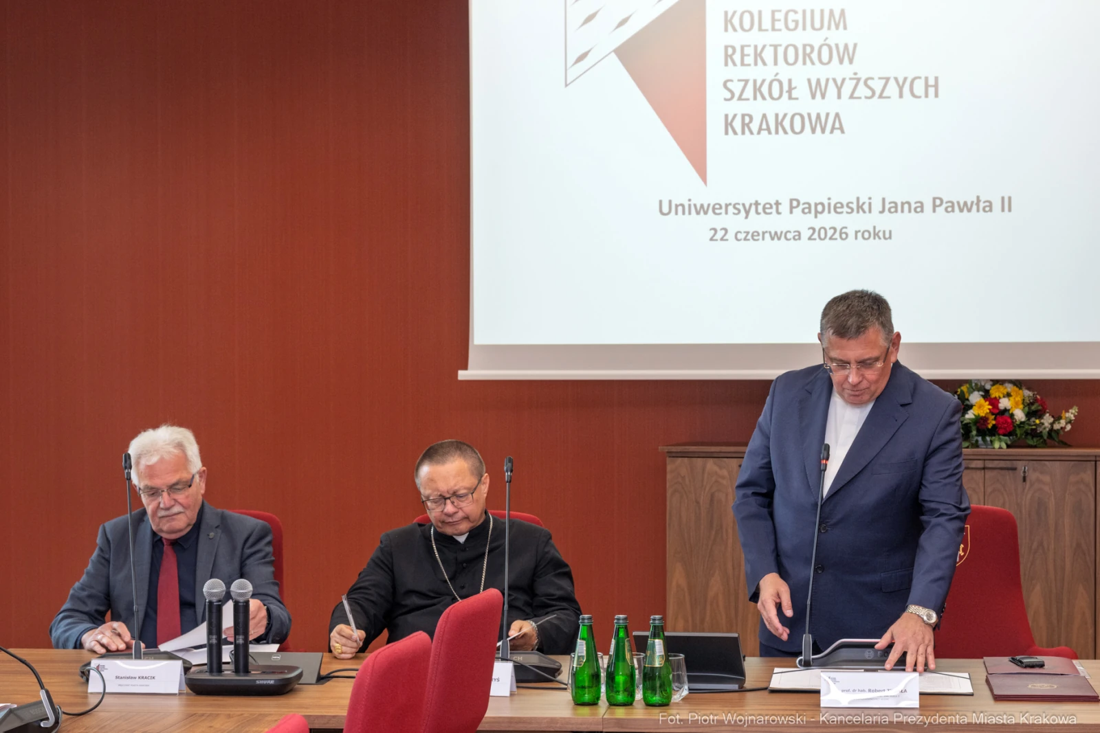 Trzech mężczyzn siedzących przy stole, w tle logo Uniwersytetu Papieskiego Jana Pawła II.  Autor: Piotr Wojnarowski