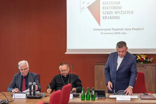 Trzech mężczyzn siedzących przy stole, w tle logo Uniwersytetu Papieskiego Jana Pawła II.