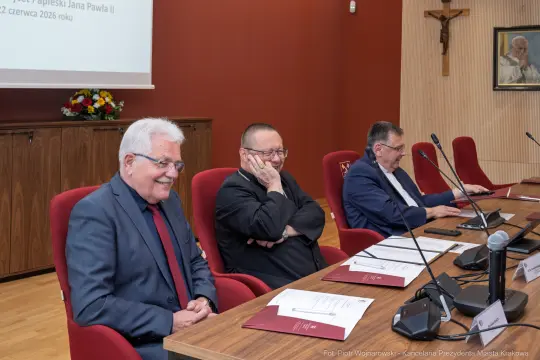 Trzech mężczyzn siedzących przy stole w sali konferencyjnej, z krzyżem na ścianie.