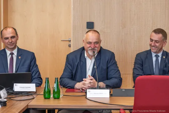 Trzech mężczyzn siedzących przy stole, rozmawiających podczas spotkania. Na stole butelki wody.