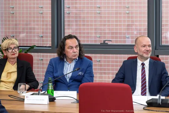 Trzy osoby siedzą przy stole, rozmawiając, w tle okna i roślinność.