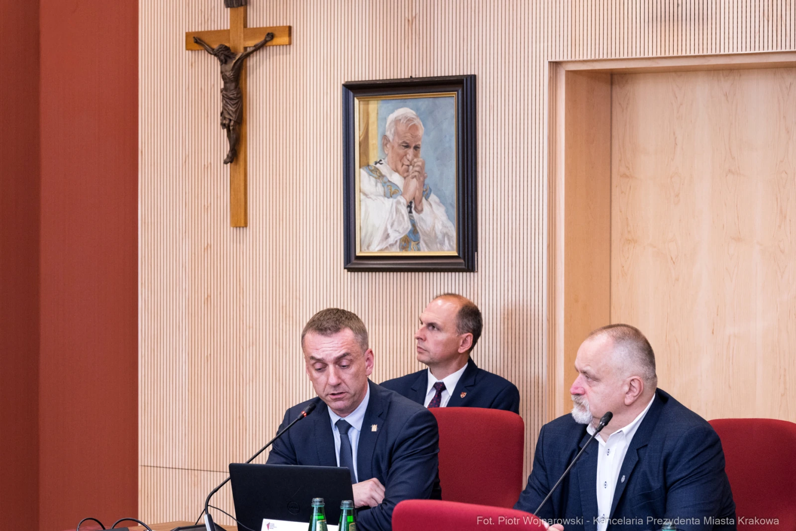Mężczyźni w garniturach podczas spotkania, w tle obraz papieża Jana Pawła II.  Autor: Piotr Wojnarowski
