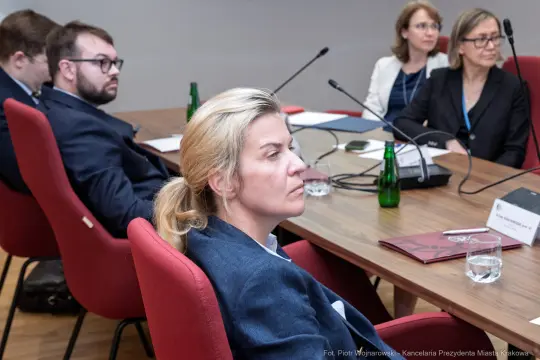 Kobieta w garniturze siedzi przy stole w trakcie spotkania, w tle inni uczestnicy.