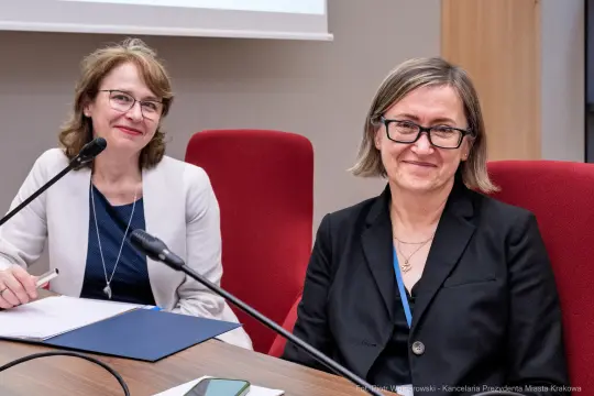 Dwie kobiety siedzą przy stole, uśmiechając się, z mikrofonami przed sobą.